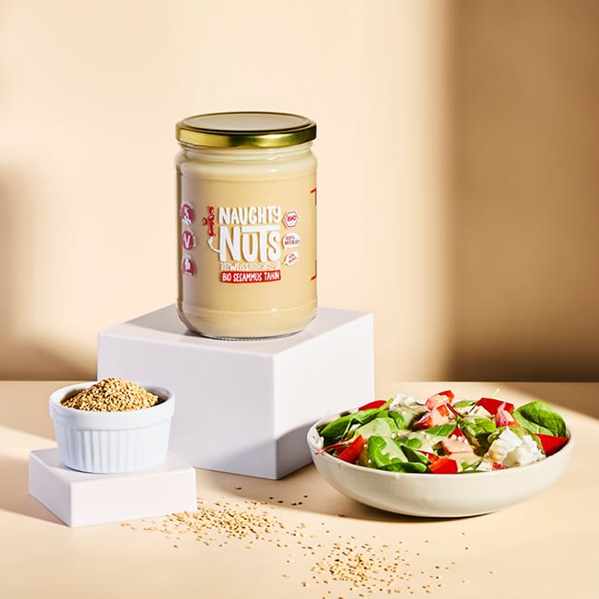 Bio Sesammus Tahin weiß (500g Glas) auf weißem Podest, daneben Schale mit Sesamsamen und Salat